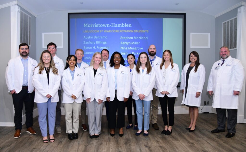 Eight women and six men standing and smiling in front of a screen titled Morristown-Hamblen. All are wearing white coats.