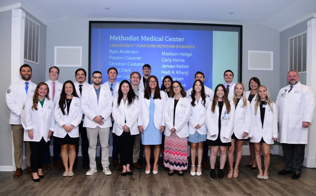 Eleven women and 10 men standing and smiling in front of a screen titled Methodist Medical Center LMU-DCOM. All are wearing white coats.