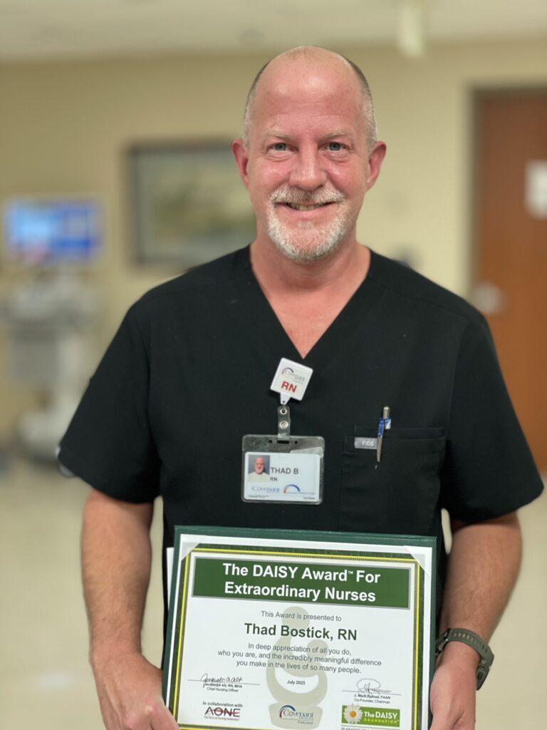 Thad Bostick, a male nurse with gray facial hair, in black scrubs, smiling and holding DAISY certificate.