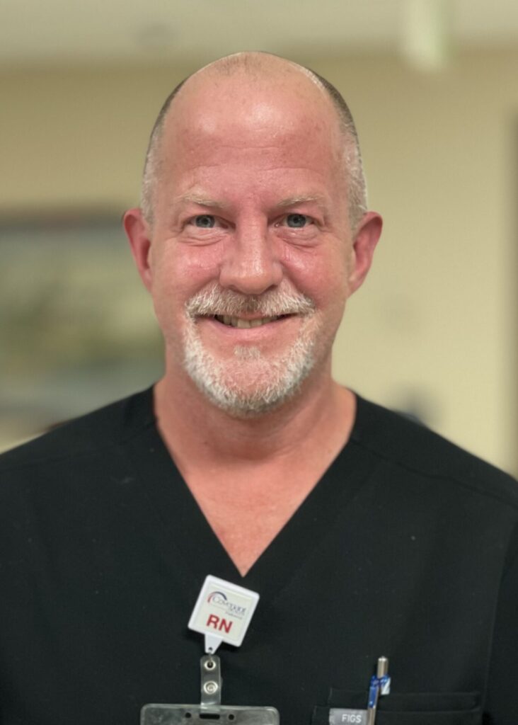 Male nurse in black scrubs, gray facial hair, smiling.