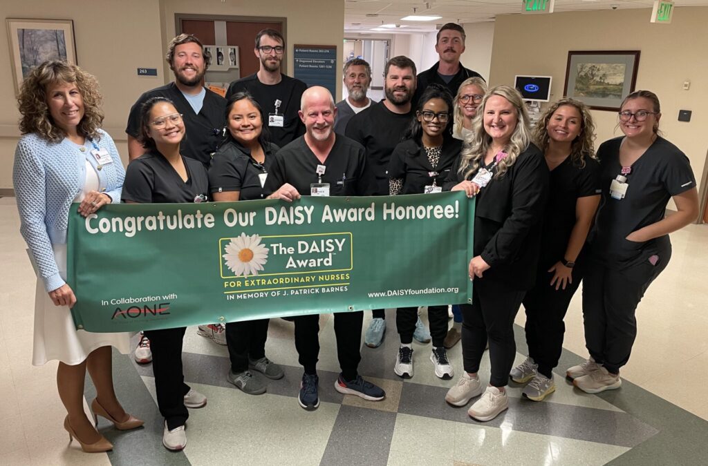 Group of nurses in black scrubs, smiling, holding a DAISY Award banner. Thad Bostick is in front row center.