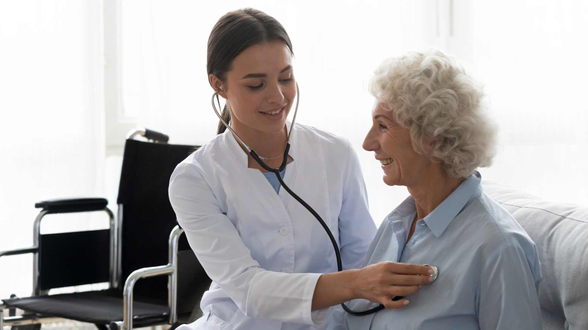 A young female doctor uses a stethoscope to listen to the chest of an older woman, who is smiling. They are both indoors, and a wheelchair is visible in the background.