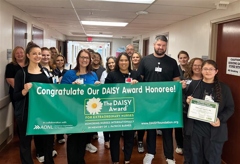 A group of nurses and staff stand in a hallway holding a green banner that reads, Congratulate Our DAISY Award Honoree! One person holds a certificate, celebrating an award for extraordinary nurses.