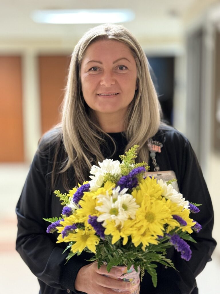 Headshot of blond nurse, holding arrangement of daisies.