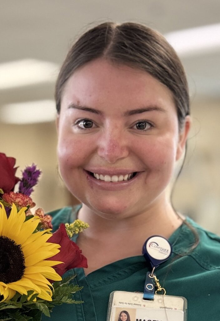 A smiling woman in green scrubs holds a colorful bouquet featuring a sunflower. Wearing a name badge and standing in a softly blurred indoor setting, she proudly celebrates her Macey Scott BEE Award.