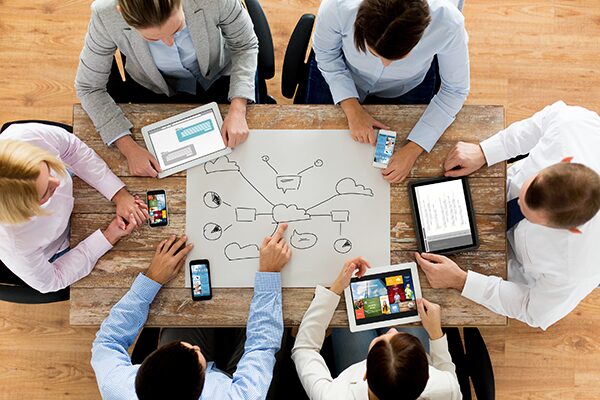 Six people sit around a wooden table, collaborating on a cloud computing diagram drawn on paper. They use tablets, smartphones, and a laptop, with documents and graphics visible on their screens.