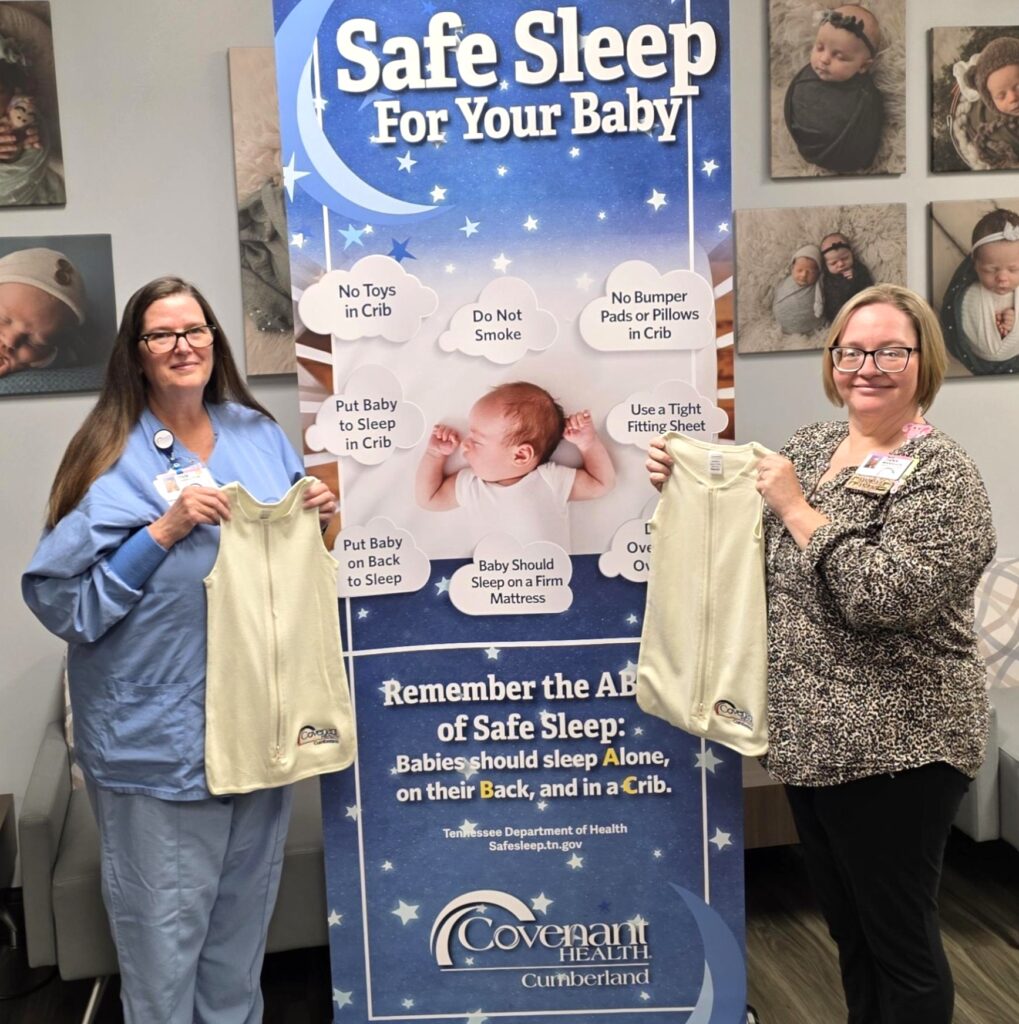 Two women stand on either side of a Safe Sleep For Your Baby banner, each holding a baby sleep sack. The banner displays safe sleep tips with images of babies in cribs. Photos of babies are on the wall behind them.
