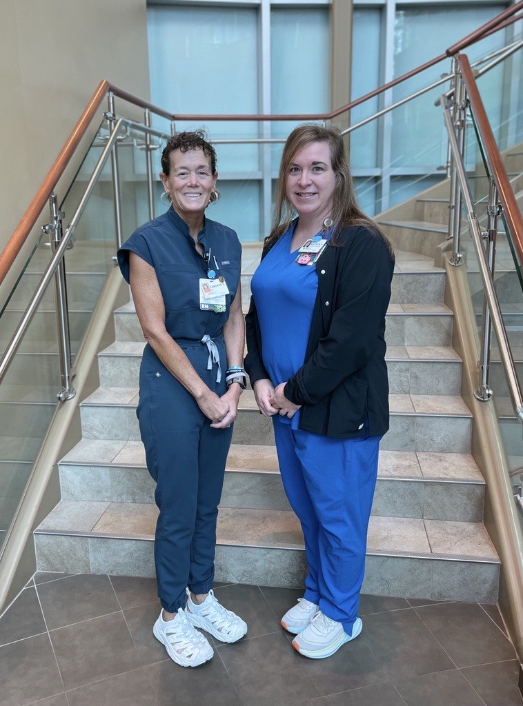 Two women in medical scrubs stand side by side, smiling, at the bottom of a staircase with glass railings in a well-lit indoor setting at Fort Sanders Regional Stroke Center. Both wear ID badges and white sneakers.