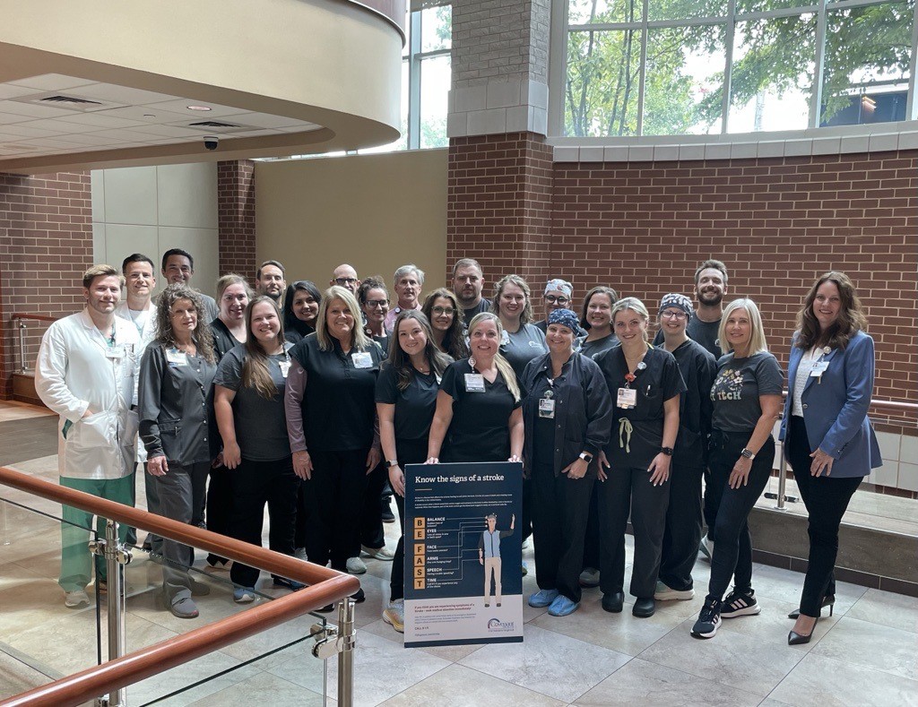 A diverse group of healthcare professionals pose together indoors near a large window and brick wall, smiling at the camera. In front of them is a sign about Fort Sanders Regional stroke awareness.