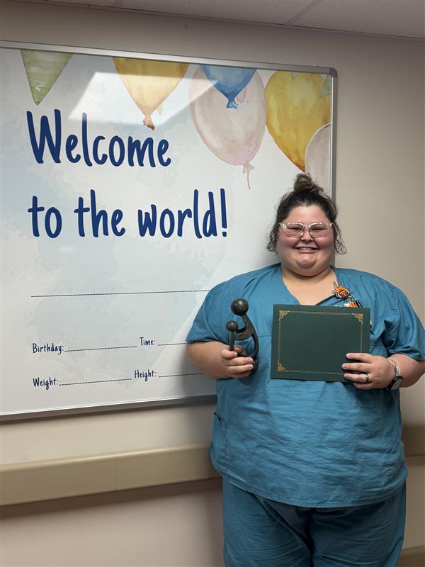 A smiling Katherine Lagunas RN, recently honored with the DAISY Award, stands in blue scrubs beside a Welcome to the world! sign, holding a green certificate and a statue shaped like a parent holding a child. The sign includes spaces for baby details like birthday and weight.