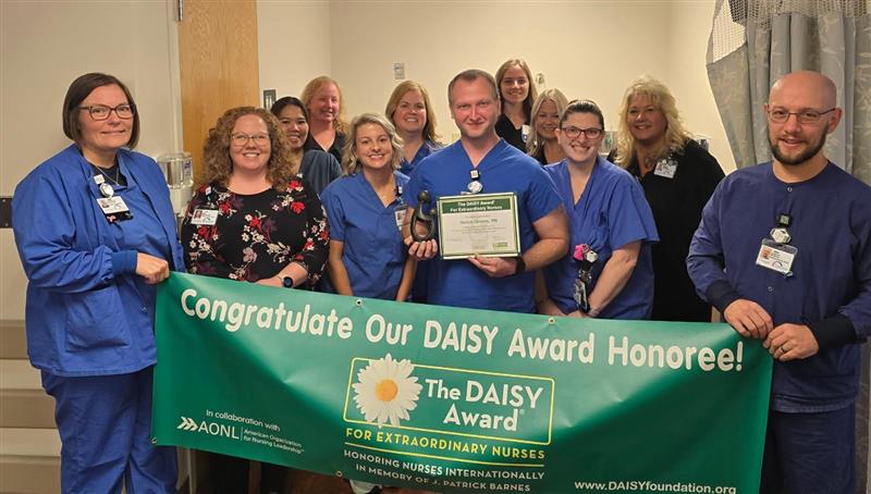 A group of nurses and staff smiling indoors, holding a green banner that reads “Congratulations Our DAISY Award Honoree!” 