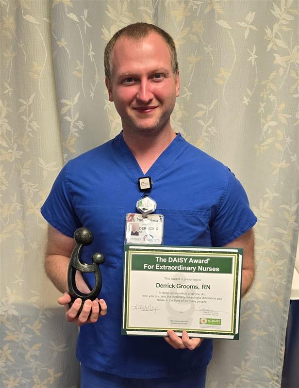 A nurse in blue scrubs holds a black trophy and a certificate that reads The DAISY Award for Extraordinary Nurses. He is smiling, standing in front of a light-colored curtain, celebrating as Lori Matthews RN Honored with DAISY Award.