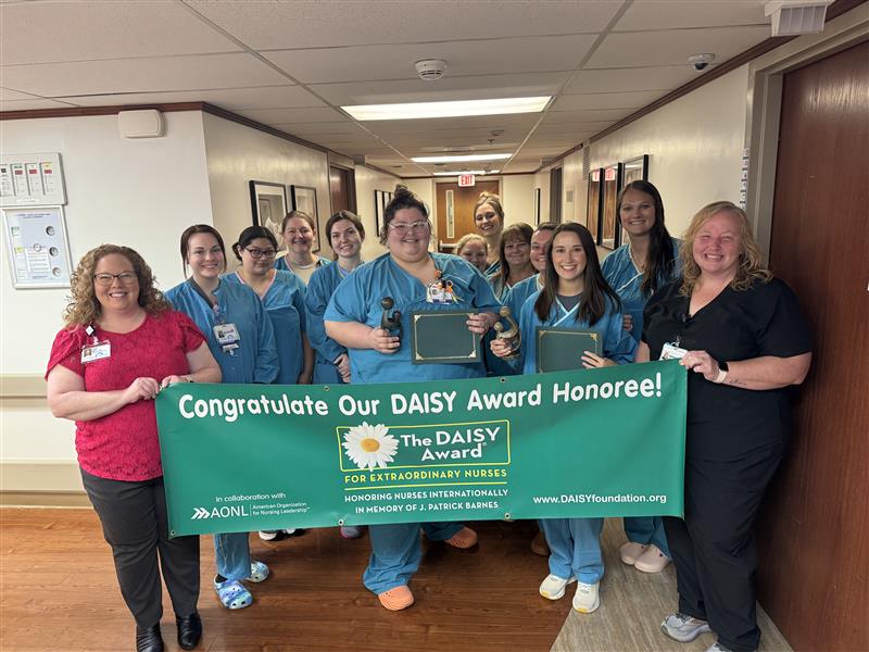 A group of nurses and staff stand in a hospital hallway holding a green banner that reads, “Congratulate Our DAISY Award Honoree!” as they celebrate Katherine Lagunas RN honored with the DAISY Award, proudly smiling and displaying certificates.