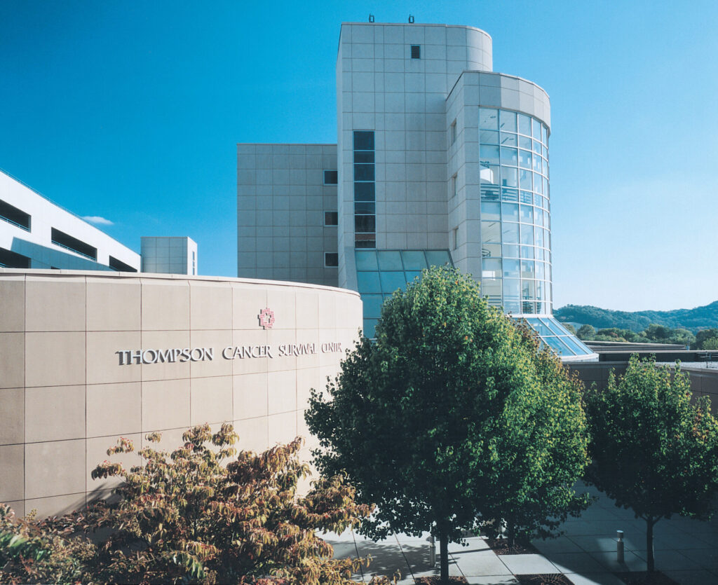 A modern building with large windows and a curved wall labeled Thompson Cancer Survival Center, surrounded by green trees under a clear blue sky.