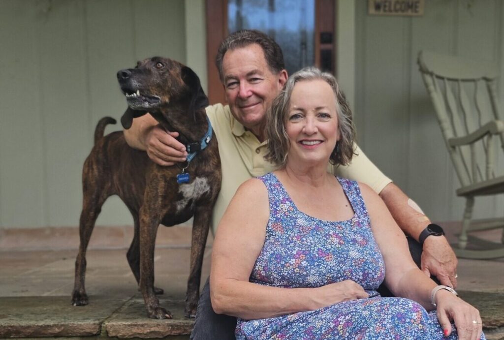 A smiling older couple and their dog sit together on stone steps outside a house