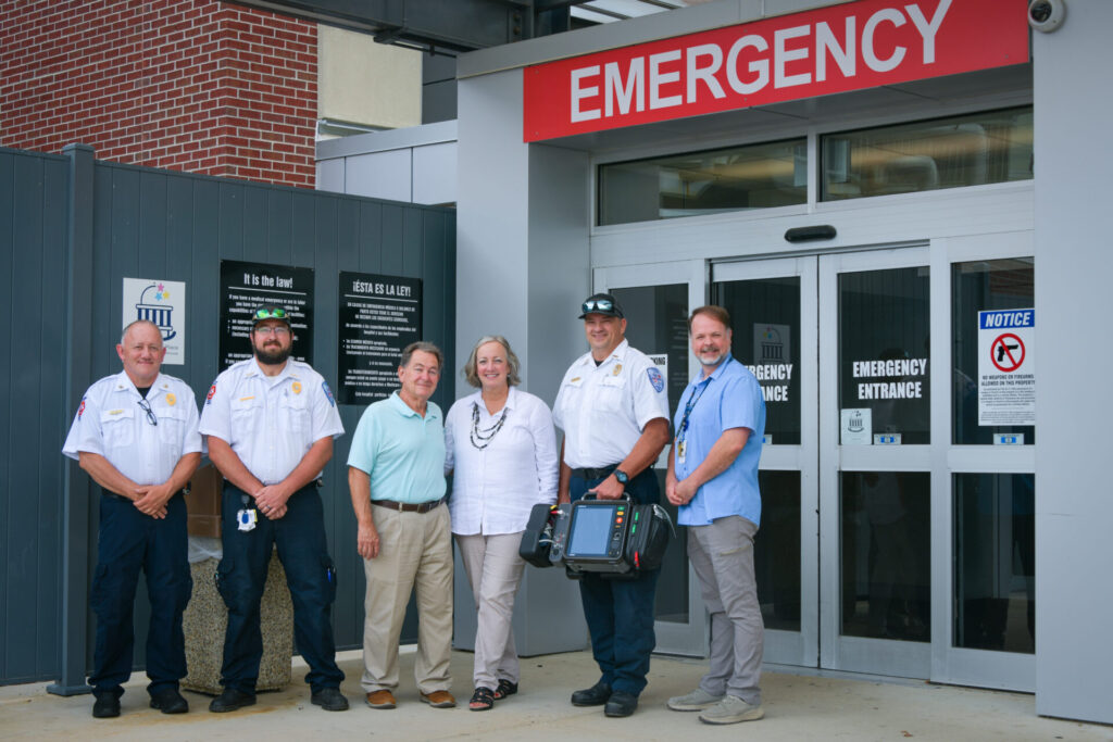 Four medical professionals, an older male patient, and his wife stand smiling outside a hospital emergency entrance under a red EMERGENCY sign. One person holds medical equipment.