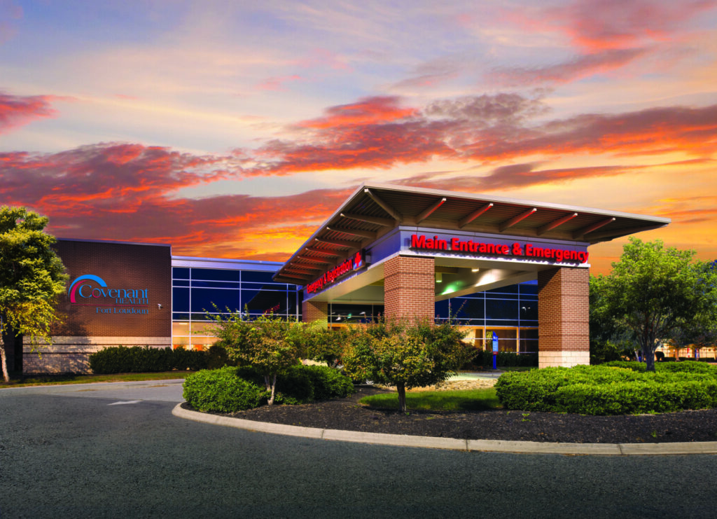 A hospital building with a lit “Main Entrance & Emergency” sign at sunset. The modern exterior has large windows, landscaping, and the Covenant HealthCare logo on the left side of the building.