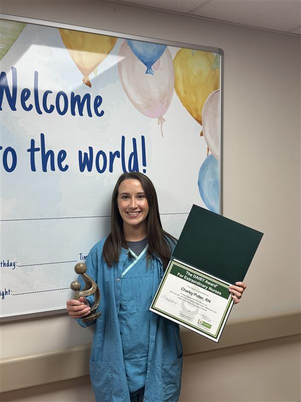 RN, Honored with DAISY Award, smiles in scrubs while holding a certificate and trophy. She stands before a Welcome to the world! wall sign with colorful balloon decorations.