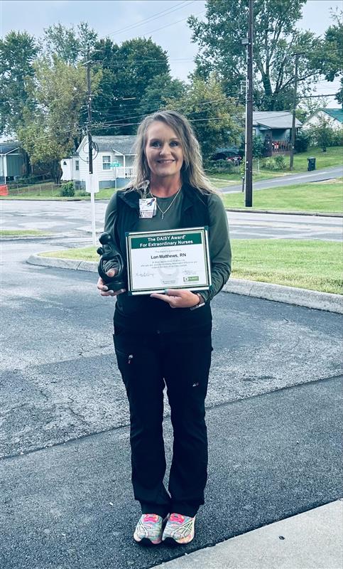 Charley Porter RN, honored with the DAISY Award, stands outdoors on a paved area holding her award certificate and a small sculpted figure. She is smiling in a green shirt and black pants, with houses and trees in the background.