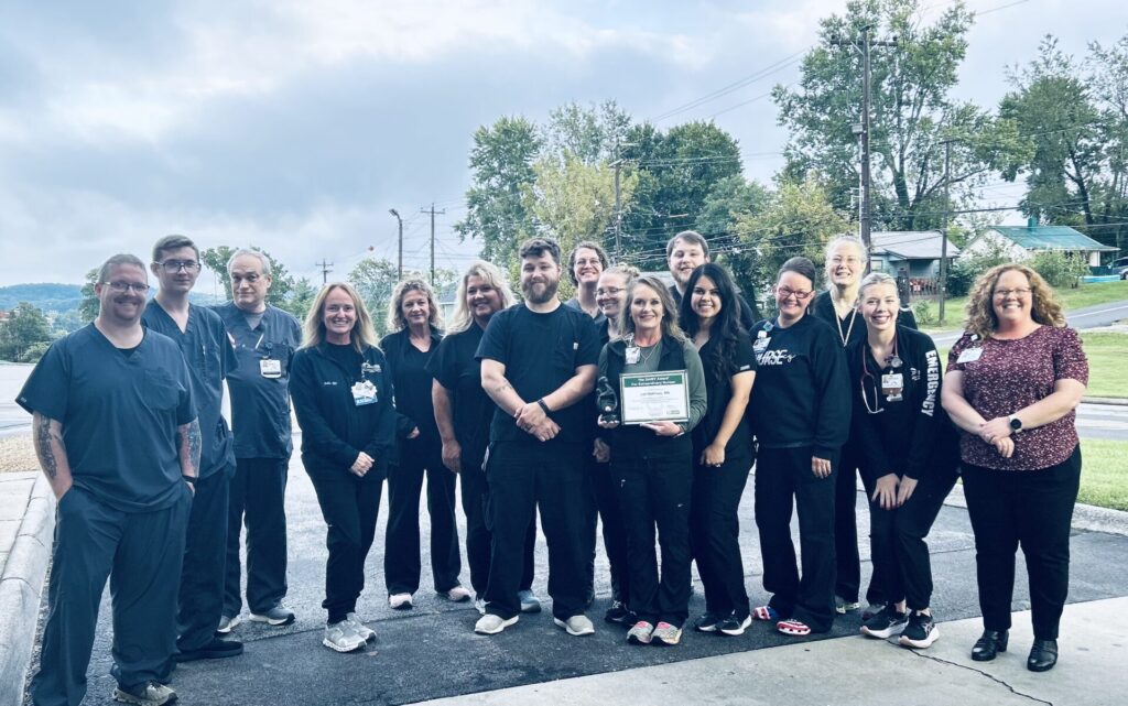 A group of healthcare workers in dark scrubs stand outside on a paved road, smiling at the camera. Charley Porter RN is near the center, proudly holding a framed certificate after being honored with the DAISY Award. Trees and houses are visible in the background.