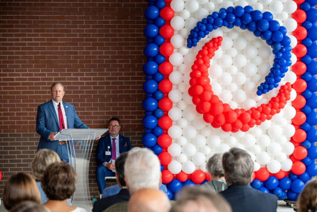 A man in a suit speaks at a podium to an audience, with another man seated nearby. Behind them is a large balloon display in red, white, and blue, forming a swirling pattern, against a brick wall.