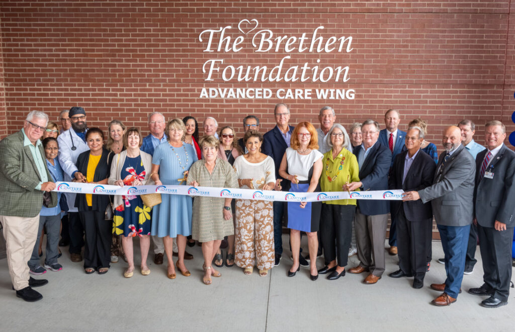 A group of people stands in front of a brick wall under a sign reading “The Brethen Foundation Advanced Care Wing,” holding a ribbon for a ceremonial cutting. Most are smiling and dressed in business or semi-formal attire.