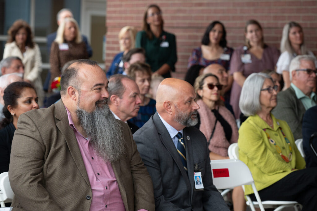 A group of people sit attentively at an indoor event. Two men in suits with beards are in the foreground, while other attendees, both men and women, are seated and standing in the background.
