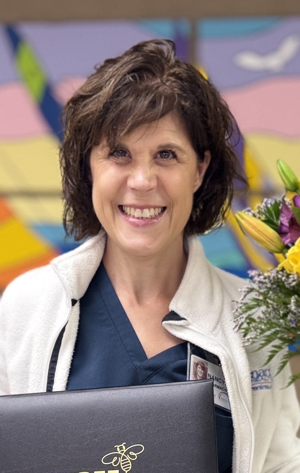 A woman with short brown hair smiles while holding a bouquet of flowers and a black folder with a gold bee emblem. She wears a navy scrub top and white jacket. A colorful abstract mural is in the background.