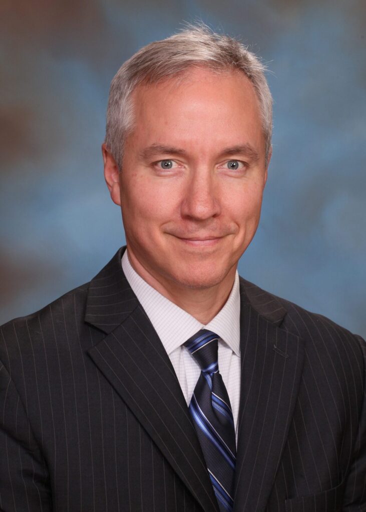 A middle-aged man with short gray hair wearing a dark pinstripe suit, light shirt, and striped tie, smiling slightly, poses in front of a blurred blue and brown background.