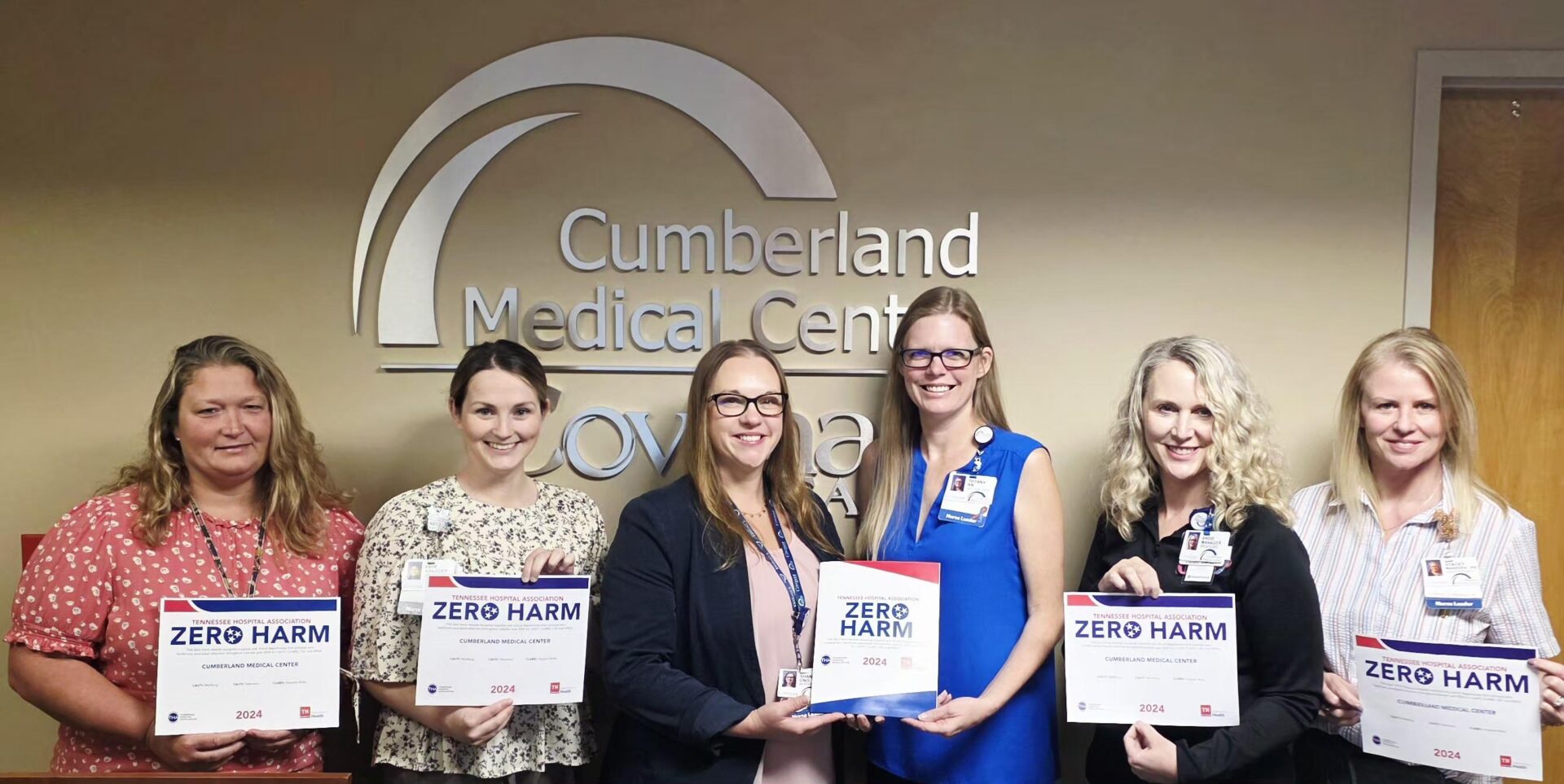 Six women stand in front of a Cumberland Medical Center sign, smiling and holding Zero Harm certificates and folders, indicating an award or recognition event.