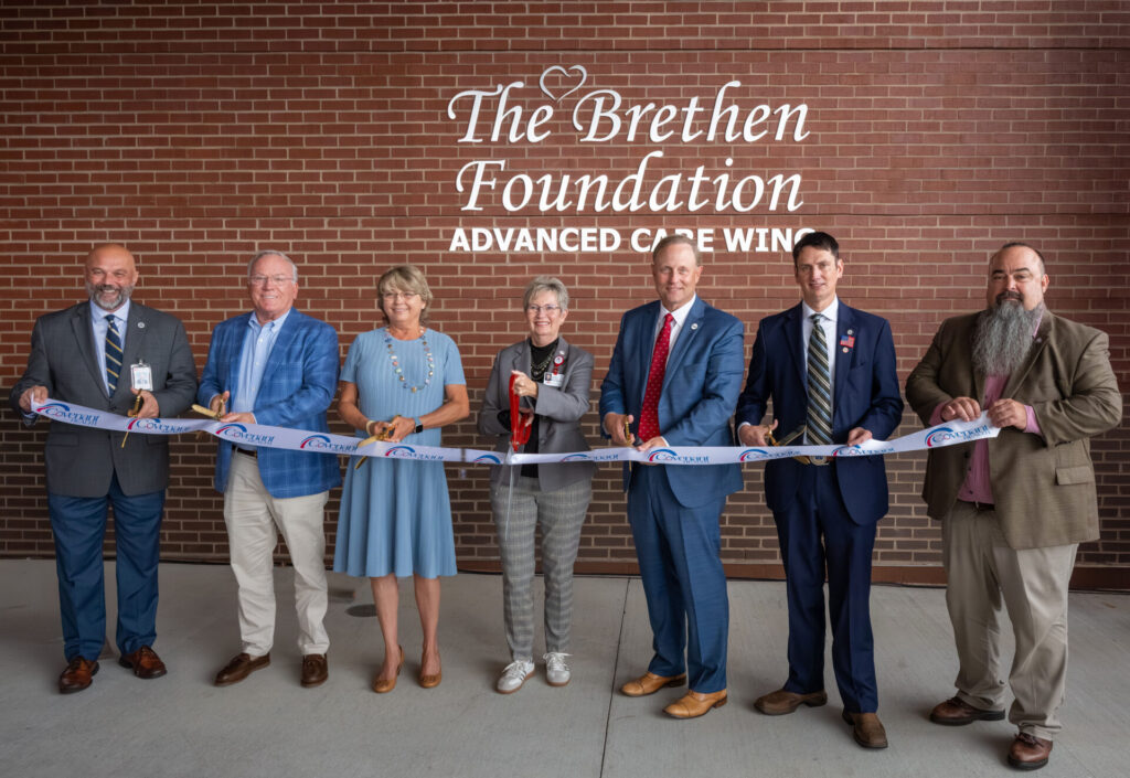 Seven people stand in a row, smiling and holding a ceremonial ribbon in front of a brick wall that reads The Brethen Foundation Advanced Care Wing. Some hold large scissors, indicating a ribbon-cutting event.