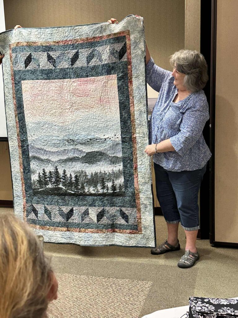 A woman stands indoors holding up a quilt featuring a scenic landscape with mountains, trees, and bird designs—a creative gift she made during her time at the Methodist Family Medicine Residency. She wears a blue patterned shirt and capri pants.