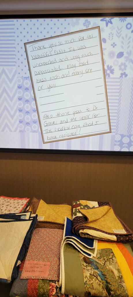 A handwritten thank-you note is projected on a screen above a table displaying several folded, colorful quilts. The note expresses gratitude for the quilts and the care received from the Methodist Family Medicine Residency team.