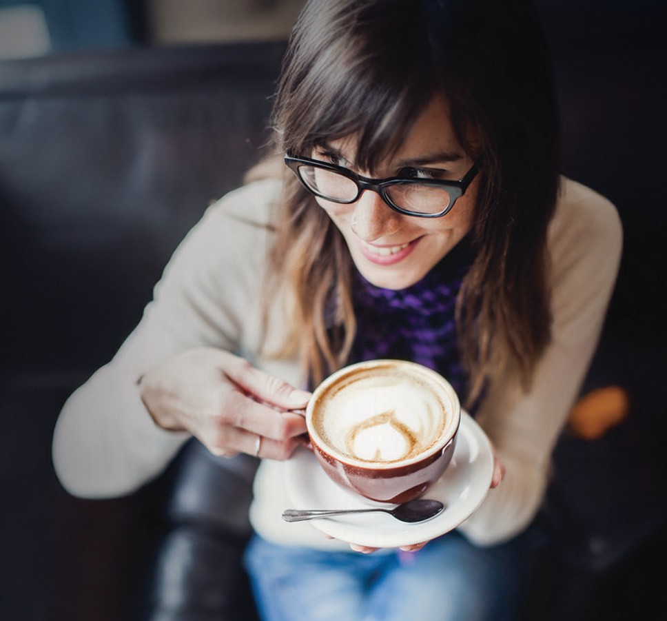 0813-5042_invite copy A woman with brown hair and glasses smiles while enjoying a cup of coffee with latte art, taking a peaceful break from the hormone roller coaster of menopause as she sits on a dark couch. A spoon rests on the saucer.
