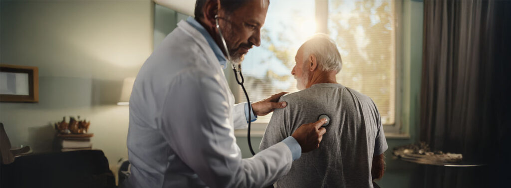 A doctor uses a stethoscope to listen to the back of an older male patient in a well-lit medical exam room with large windows and curtains.