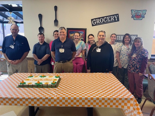 A group of eleven people stands behind a table with an orange-checkered tablecloth and a decorated cake. They are indoors, and the wall behind them has signs reading GROCERY and Salty Sea.