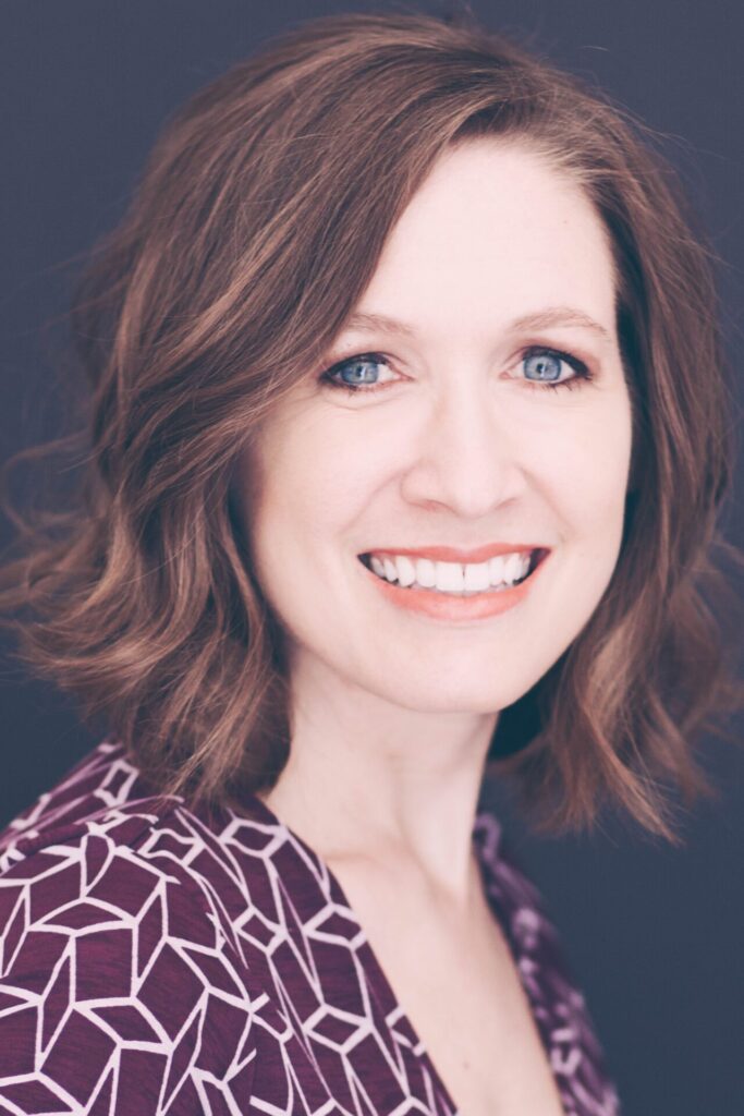 A woman with shoulder-length wavy brown hair smiles at the camera. She has blue eyes and is wearing a purple patterned top. The background is dark and plain.