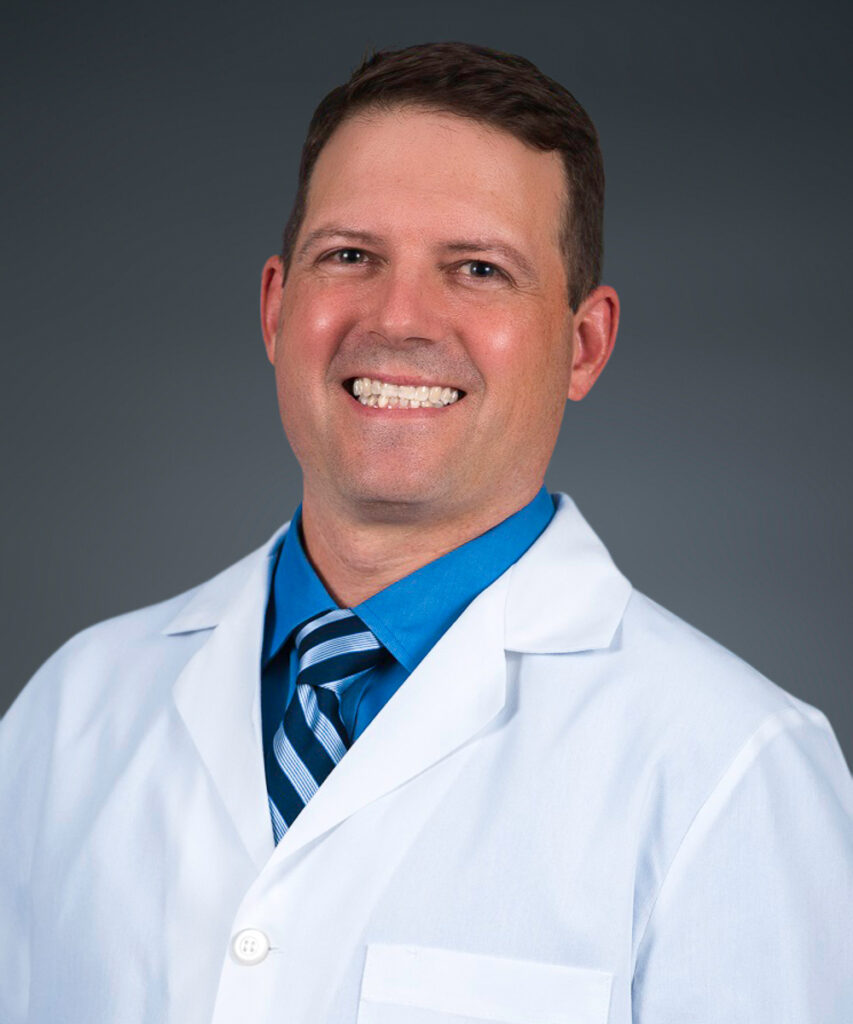 A smiling man wearing a white lab coat, blue shirt, and striped tie poses against a plain dark gray background.