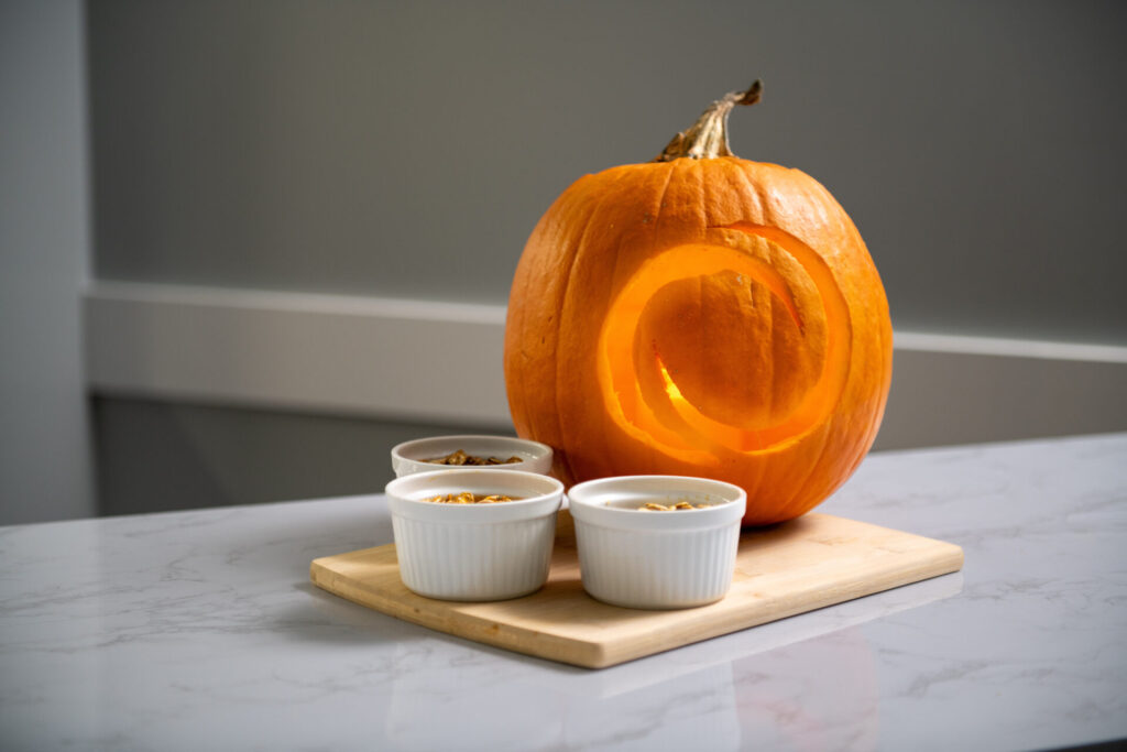 A carved pumpkin with a circular design sits on a wooden board beside three small white ramekins, each filled with roasted pumpkin seeds, on a marble countertop.