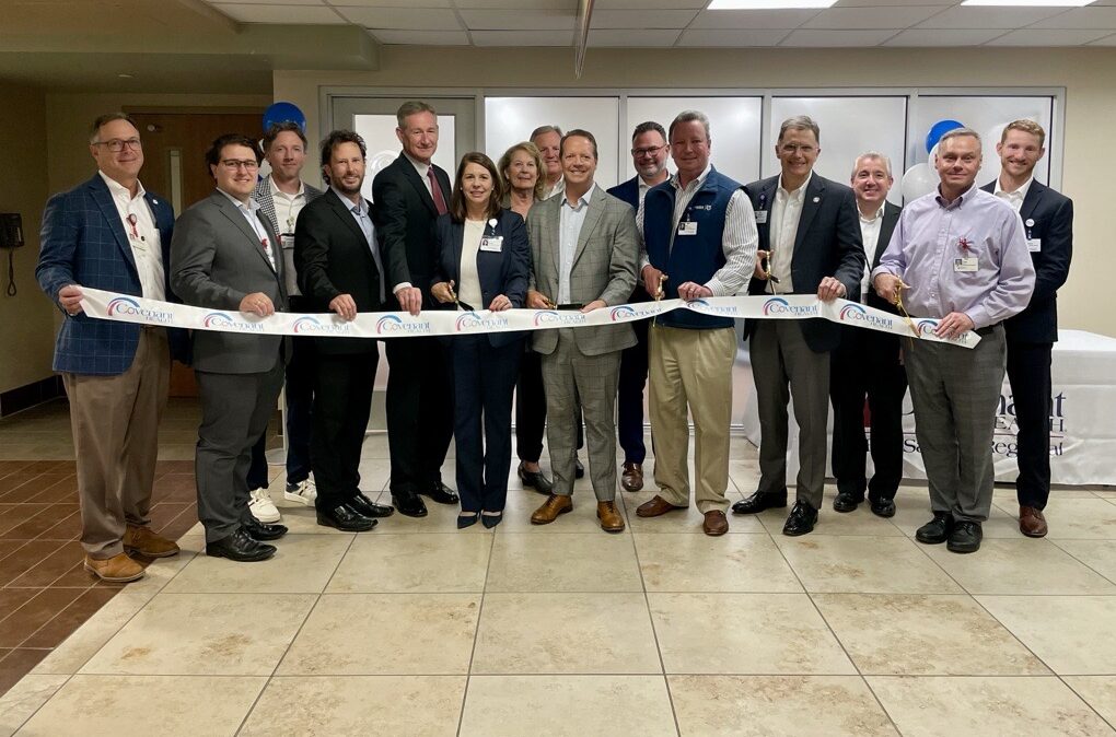 A group of people in business attire stand indoors in a semicircle, smiling and holding a ceremonial ribbon, as if celebrating a grand opening or special event.