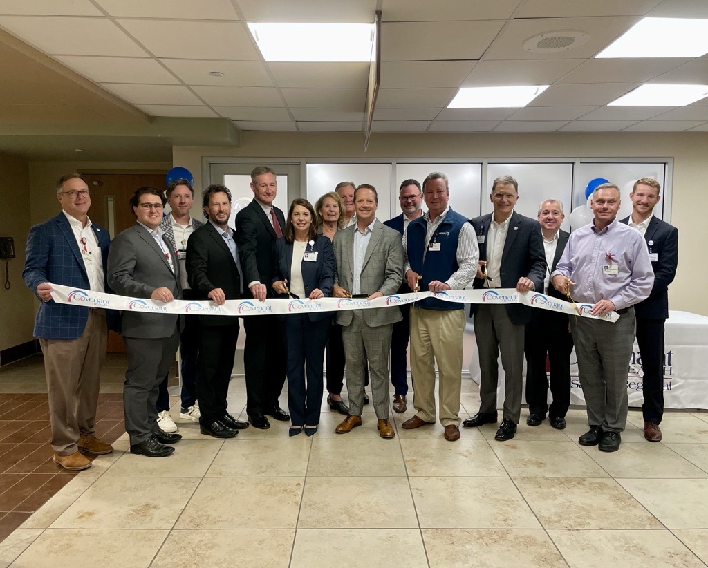 A group of people in business attire stand indoors in a semicircle, smiling and holding a ceremonial ribbon, as if celebrating a grand opening or special event.