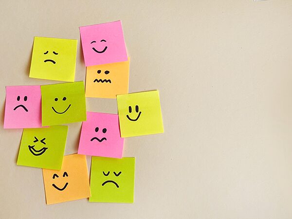 Nine sticky notes with drawn faces showing various emotions, including happy, sad, angry, and confused, are arranged on a beige surface. The notes are pink, yellow, and orange. The right side of the image is blank.