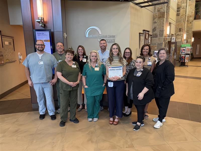 A group of twelve hospital staff members pose and smile in the lobby of LeConte Medical Center. One woman in the front center holds a certificate that reads ZERO HARM. The setting appears friendly and professional.