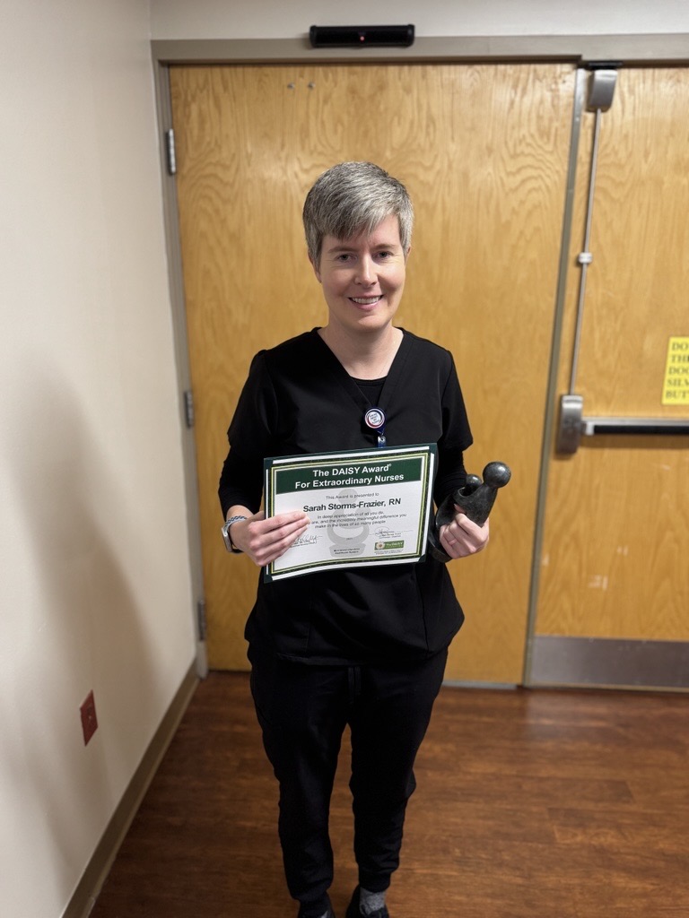 Derrick Grooms RN, Honored with DAISY Award, stands indoors in black scrubs, holding a certificate and a small black object, smiling at the camera in front of closed wooden doors.