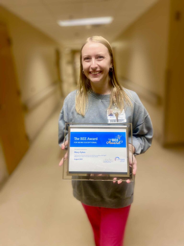 A smiling woman stands in a hallway holding a framed BEE Award certificate for being exceptional. She wears a gray sweatshirt, pink pants, and a name badge.