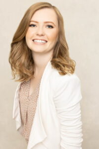 A young woman with wavy, light brown hair smiles warmly at the camera. She is wearing a white blazer over a patterned blouse, standing against a neutral, light background.
