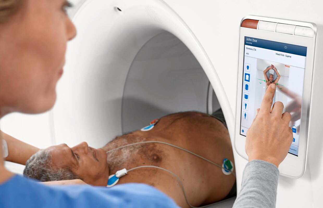 Patient in CT machine A medical professional operates a touchscreen control panel next to a patient lying inside a CT scanner with electrodes attached to his chest.
