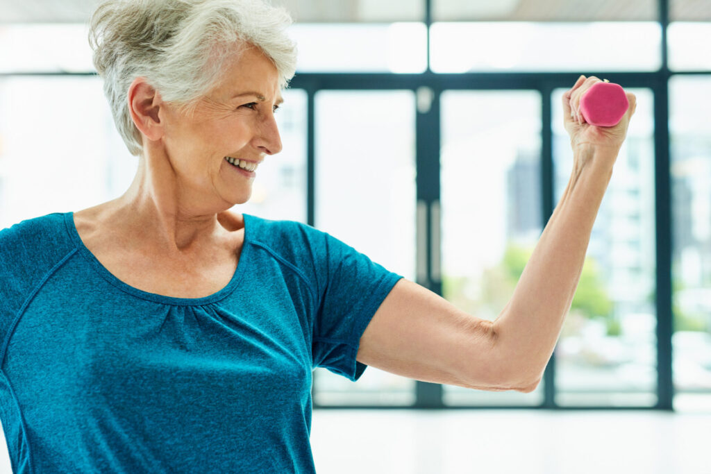 Smiling older woman with short gray hair lifts a pink dumbbell with one arm, wearing a blue shirt, standing indoors in front of large windows—showcasing how exercise can help combat depression in seniors.