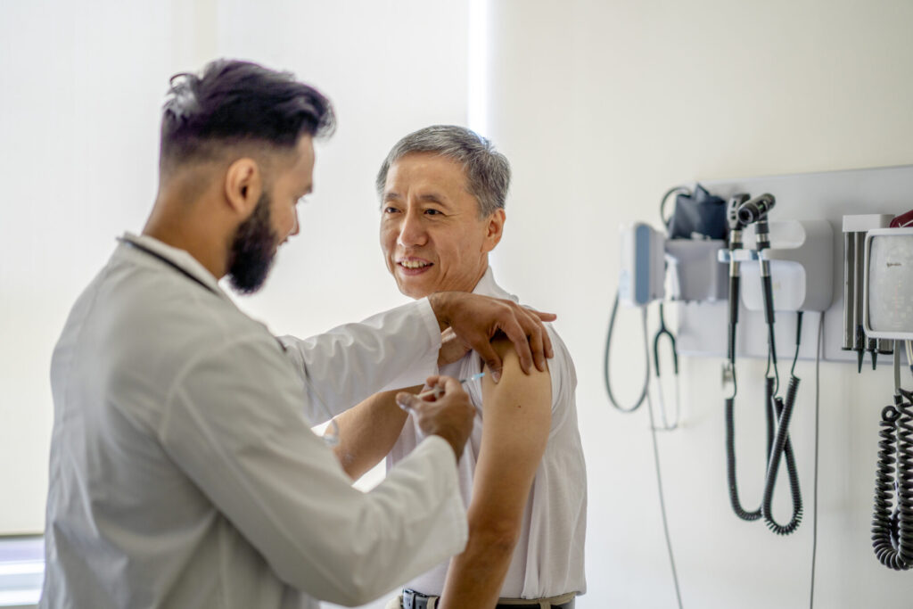 Older man sitting in a doctor's office smiling at a male doctor giving him a shot