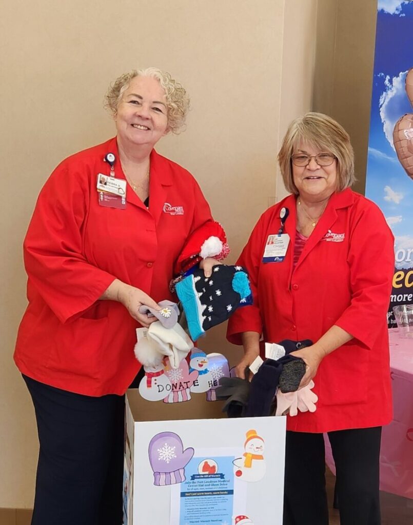 Two women in red shirts stand beside a decorated donation box filled with winter hats, gloves, and stuffed animals. Their smiles reflect why volunteering matters at Fort Loudoun Medical Center, as they help spread warmth and joy indoors.